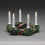A six-light Christmas wreath candle holder with green pine leaves, a red ribbon, and six white taper candles with lit flame tips, set against a dark gray studio background.