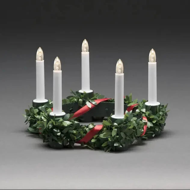 A six-light Christmas wreath candle holder with green pine leaves, a red ribbon, and six white taper candles with lit flame tips, set against a dark gray studio background.