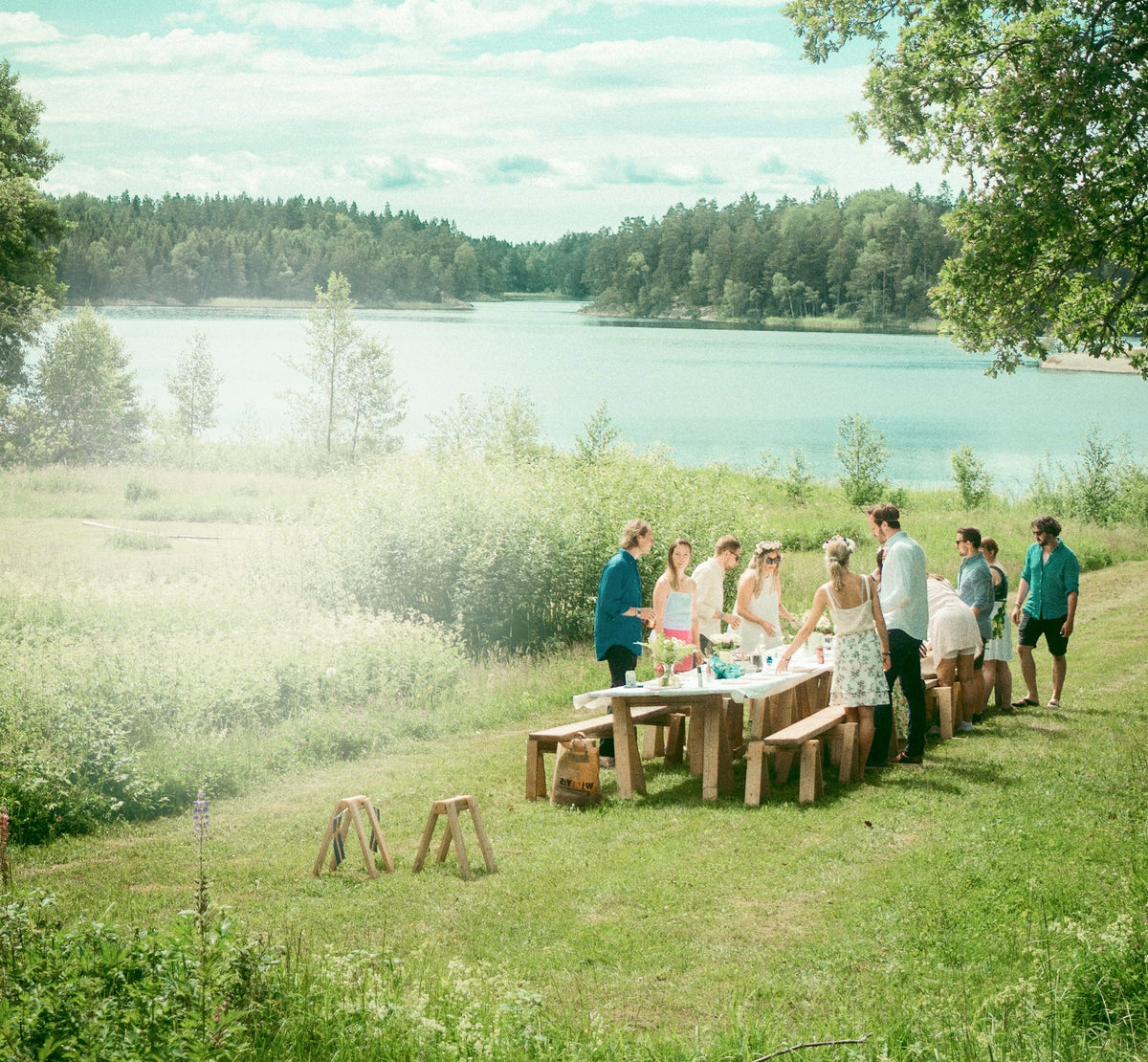 A group of people gathered around a wooden table outdoors by a lake, with grassy fields, trees, and a misty scenic background.