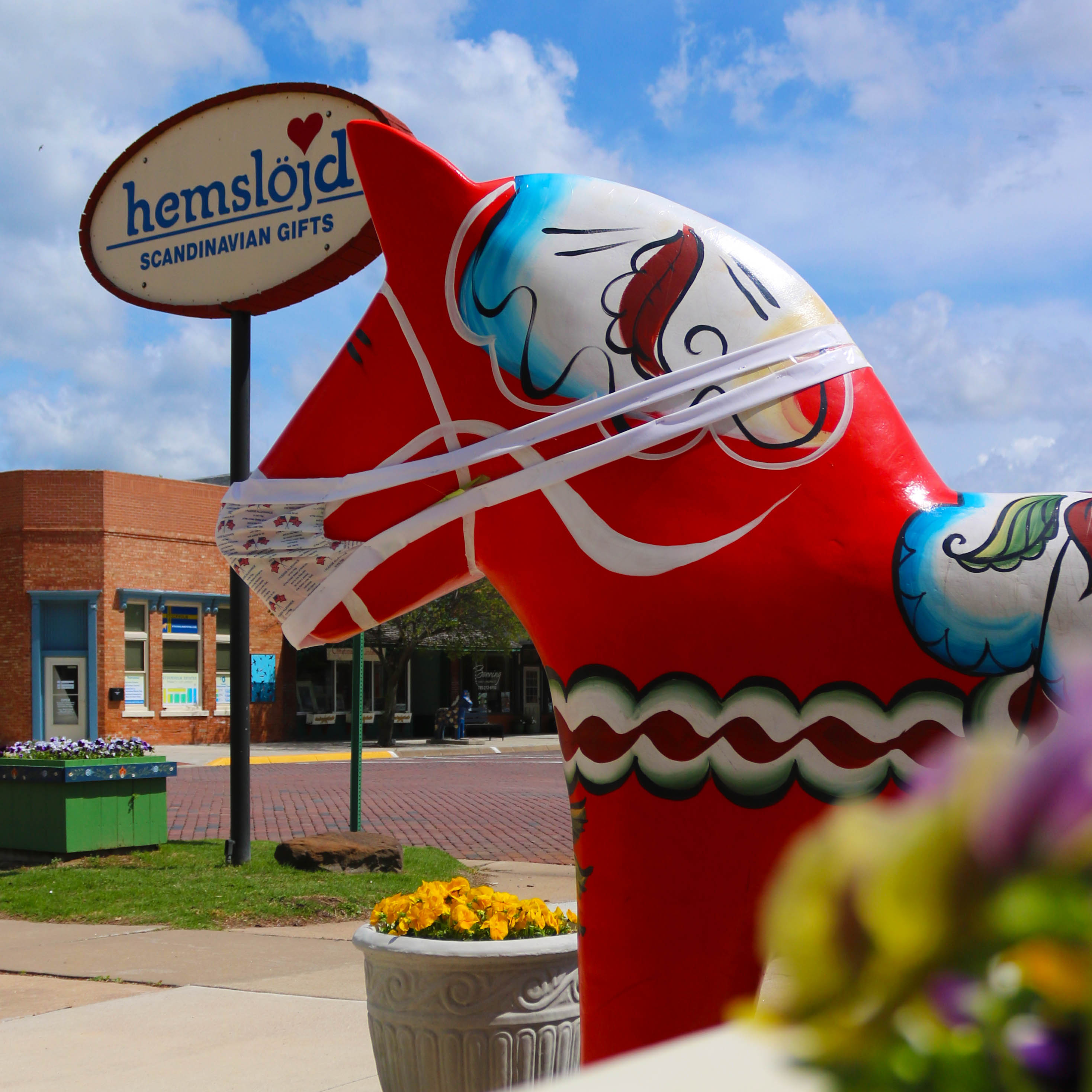 A close-up image of a red, decoratively painted Dala Horse figure with the Hemslojd storefront in the background