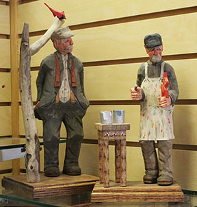 Bruce Harding's wood figurines. One is a man painting a dala horse and the other is an older man with his hands in his pockets standing by a tree with a red bird in it.
